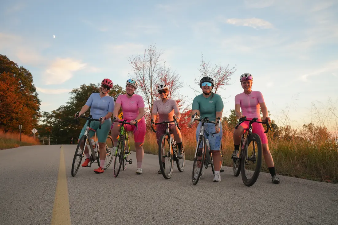 Five women in Allora kits straddling their bikes at sunset