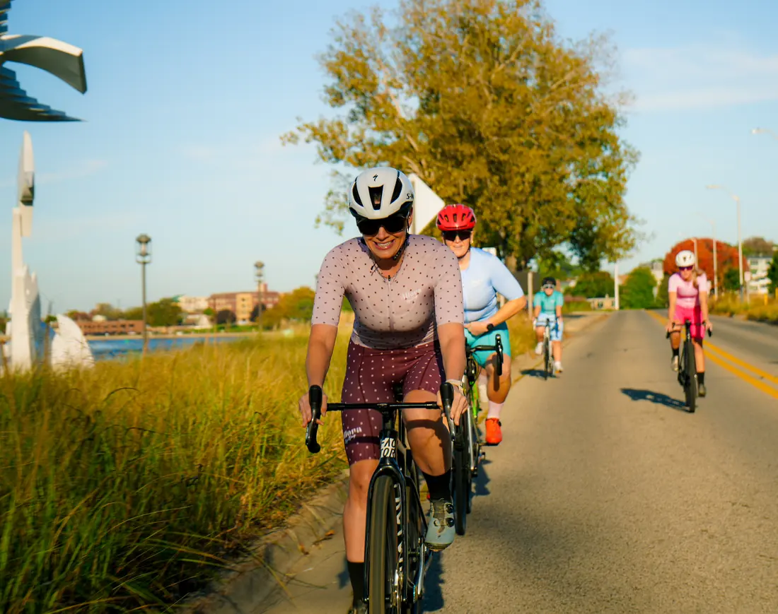 A rider in the dusty-rose kit grinning on a grassy waterfront path