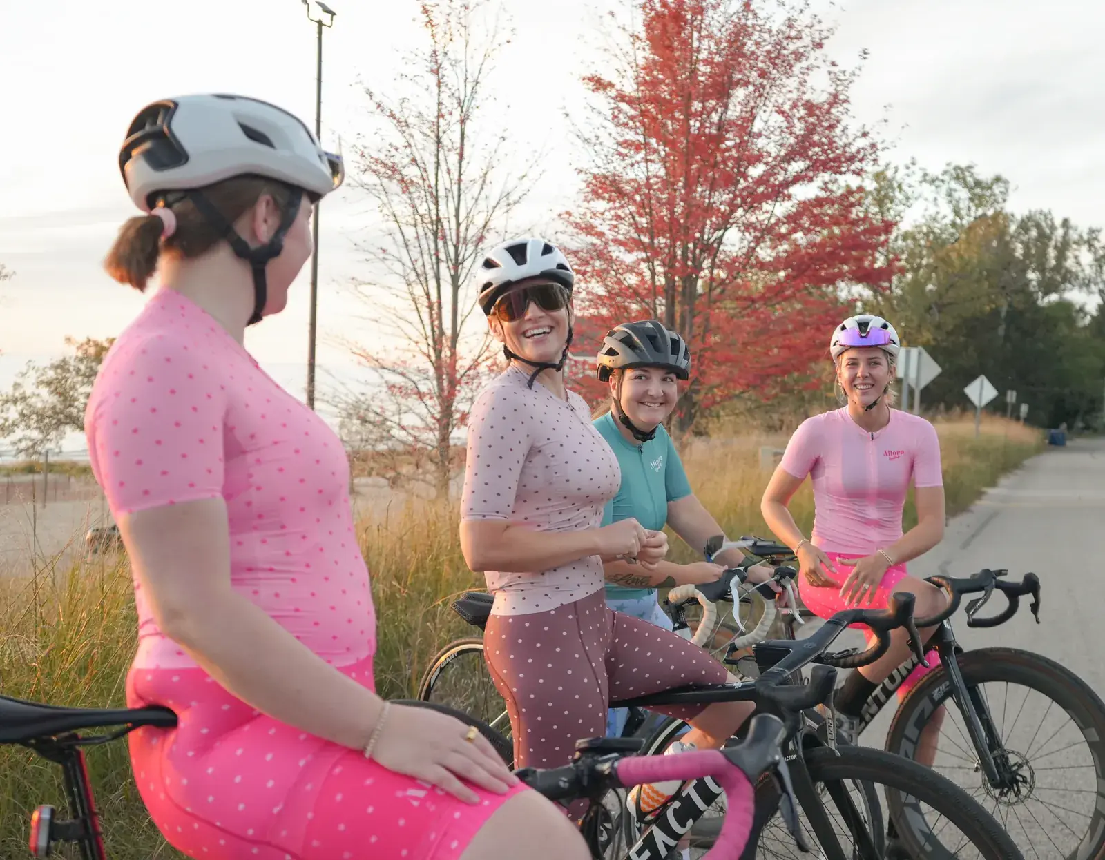Four women in Allora and other cycling kit, laughing together next to their road bikes at sunset, in pink polka-dot and teal jerseys