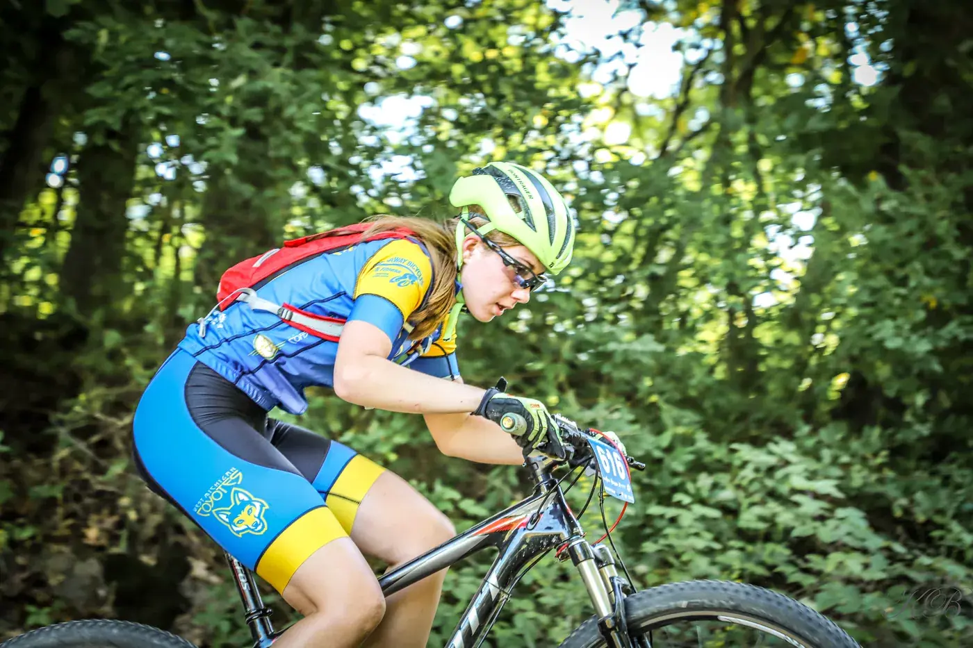 Addison racing a mountain bike through the trees in her blue and yellow West Michigan Coyotes kit